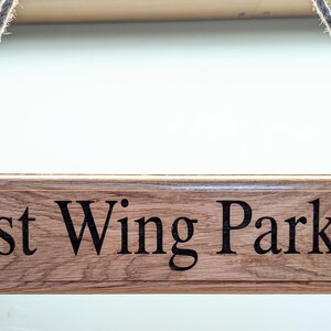 May include: A rectangular wooden sign with the words "East Wing Parking" in black lettering. The sign is suspended by a natural rope. The wood has a visible grain and a warm, brown tone.