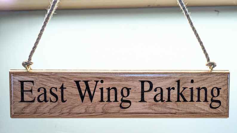 May include: A rectangular wooden sign with the words "East Wing Parking" in black lettering. The sign is suspended by a natural rope. The wood has a visible grain and a warm, brown tone.