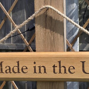 May include: Wooden sign with the text "Made in the UK" engraved in a serif font. The sign is rectangular, with a natural wood finish, and is suspended by a rope. The background includes a wooden trellis and foliage.