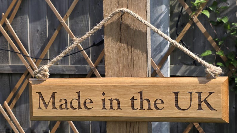 May include: Wooden sign with the text "Made in the UK" engraved in a serif font. The sign is rectangular, with a natural wood finish, and is suspended by a rope. The background includes a wooden trellis and foliage.