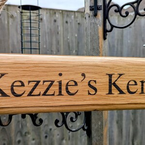 May include: A rectangular wooden sign with the text "Kezzie's Kennel" in black lettering. The sign is made of light brown wood and is suspended by a rope. The sign is likely for a dog kennel.