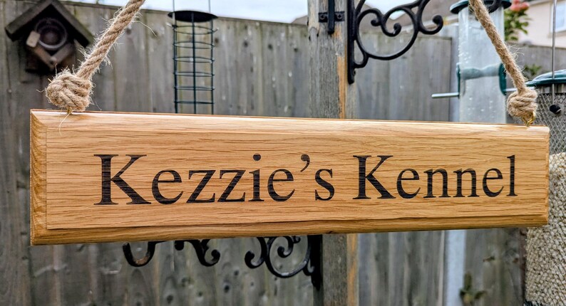 May include: A rectangular wooden sign with the text "Kezzie's Kennel" in black lettering. The sign is made of light brown wood and is suspended by a rope. The sign is likely for a dog kennel.