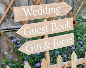 Könnte beinhalten: Drei hellbraune, pfeilförmige Holzschilder, die vertikal an einem zentralen Pfosten befestigt sind. Das oberste Schild trägt die Aufschrift Wedding, das mittlere Guest Book und das untere Gin & Tonic. Der Text ist in weißer, serifenloser Schrift.