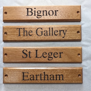 May include: Four rectangular light brown wooden signs with beveled edges and pre-drilled holes on each end. Each sign features dark, engraved text reading Bignor, The Gallery, St Leger, and Eartham, arranged in a vertical stack on a light surface.