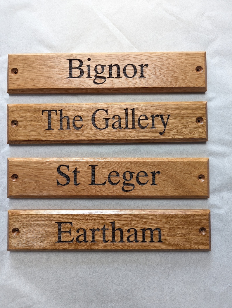 May include: Four rectangular light brown wooden signs with beveled edges and pre-drilled holes on each end. Each sign features dark, engraved text reading Bignor, The Gallery, St Leger, and Eartham, arranged in a vertical stack on a light surface.