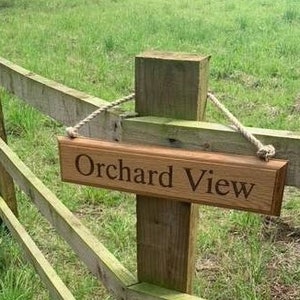 May include: A wooden sign with the words "Orchard View" engraved on it hangs from a wooden fence post. The sign is suspended by a rope. The background features a green field and a wooden fence.