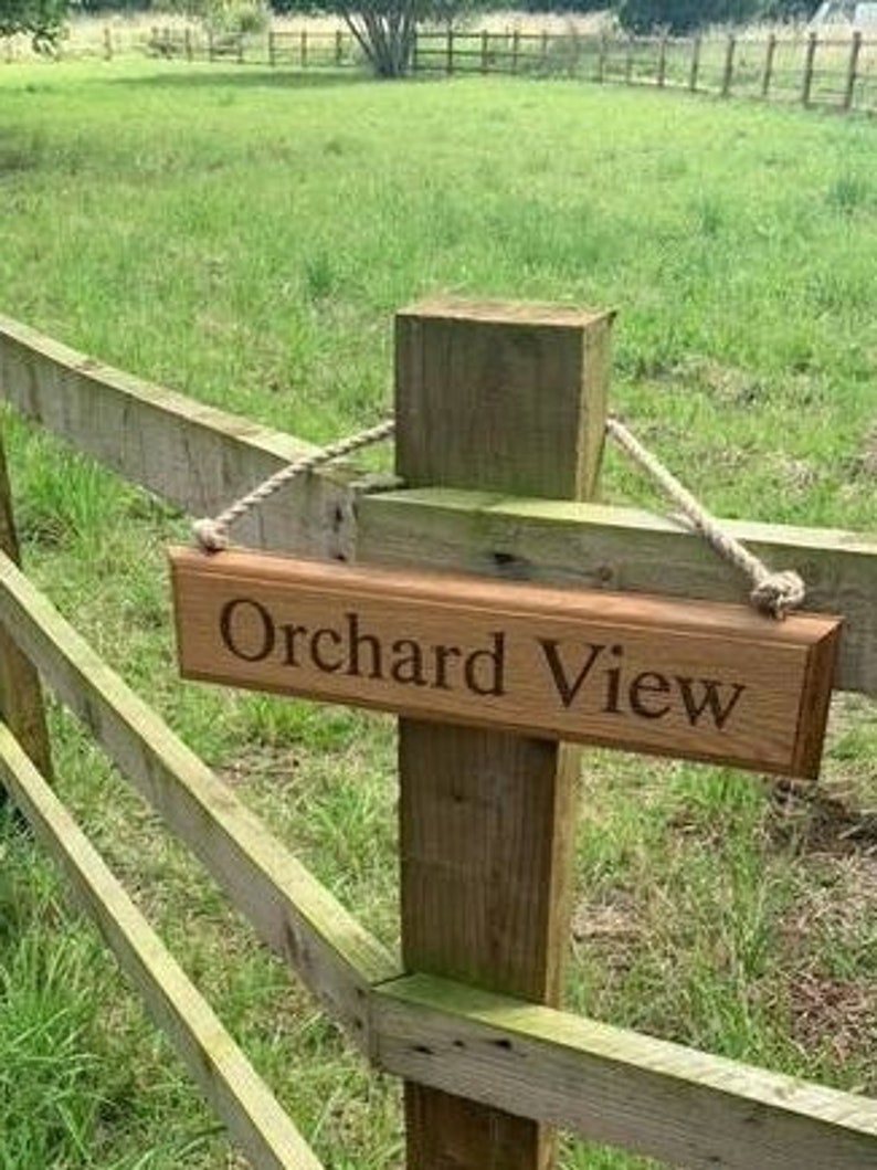 May include: A wooden sign with the words "Orchard View" engraved on it hangs from a wooden fence post. The sign is suspended by a rope. The background features a green field and a wooden fence.