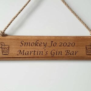 May include: Wooden sign with the text "Smokey Jo 2020 Martin's Gin Bar" engraved in a dark font. The sign has a rope for hanging and features a cocktail glass illustration on each end. The wood has a natural grain.