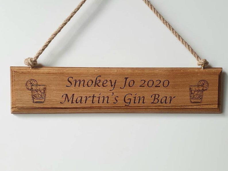 May include: Wooden sign with the text "Smokey Jo 2020 Martin's Gin Bar" engraved in a dark font. The sign has a rope for hanging and features a cocktail glass illustration on each end. The wood has a natural grain.