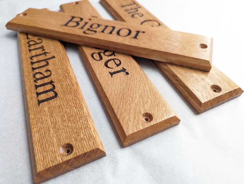 May include: Four rectangular light brown wooden signs with beveled edges and pre-drilled holes at each end. Each sign features dark, engraved text including the words Bignor, Lartham, ger, and The C, showcasing the natural wood grain texture.