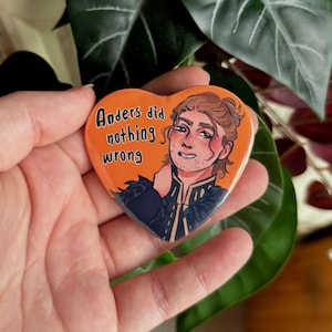 Peut inclure: Un bouton en forme de cœur avec un fond orange présente une illustration de dessin animé d'un homme aux cheveux bruns et une tenue noire et blanche. Le texte "Anders did nothing wrong" est imprimé en blanc sur le bouton.