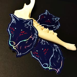 May include: Three holographic stickers of a wolf's head with red eyes and a rainbow iridescent outline on a dark blue background.