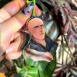 Puede incluir: Un llavero de acrílico transparente con una ilustración de dibujos animados de una persona con orejas puntiagudas, piel clara y cabello oscuro. La persona lleva una chaqueta azul oscuro con un cuello de piel marrón claro y una camisa azul oscuro con un patrón de líneas azul claro y blancas. El llavero está colgado de un anillo de plástico verde.