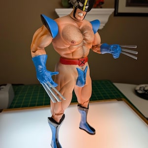 May include: A muscular action figure of a character wearing a black and yellow mask, blue shoulder pads, blue gloves with three silver claws on each hand, a red belt with an X buckle, and blue boots. The figure has a tan skin tone and chest hair.