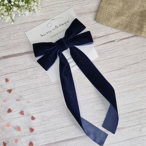 May include: A navy blue velvet bow with long tails. The bow is handmade and is on a white card with the text "bow sew cute designs handmade hair accessories".