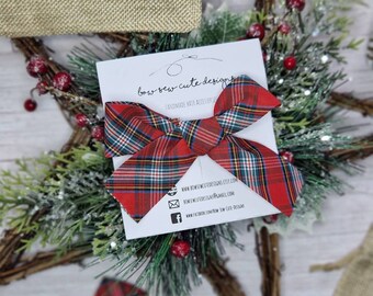 May include: A red plaid bow hair accessory is the focal point, set against a backdrop of festive greenery and a rustic star-shaped wreath. A small burlap bag with the words "handmade with love" is also visible.