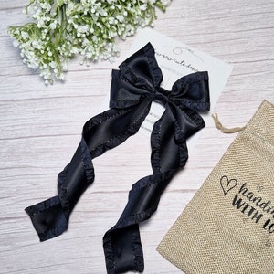 May include: Black satin ribbon hair bow with ruffled edges. The bow is tied in a large loop with two long tails. The bow is on a white wooden surface with a small white flower bouquet and a burlap bag with the text "handmade with love" printed on it.