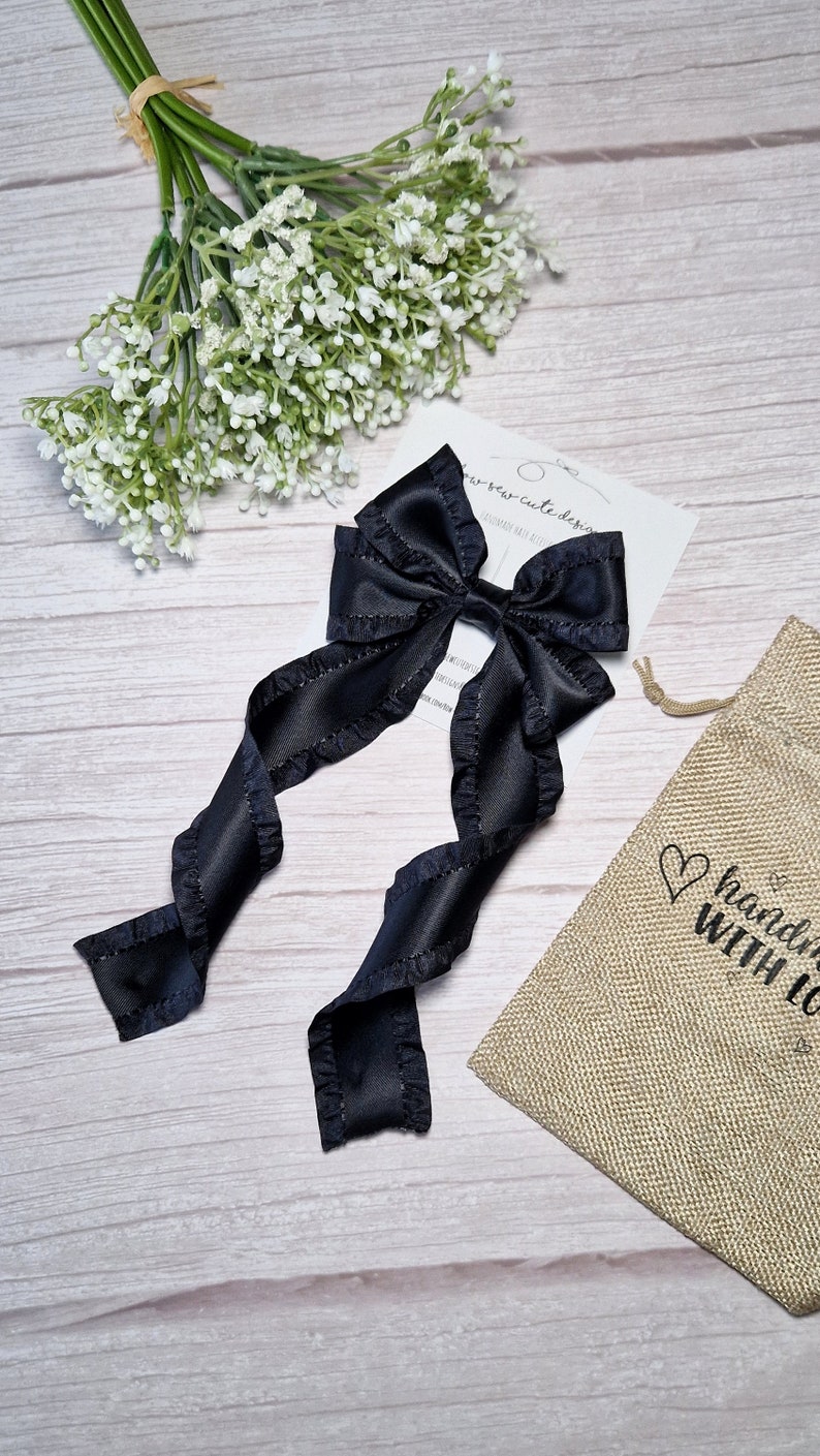 May include: Black satin ribbon hair bow with ruffled edges. The bow is tied in a large loop with two long tails. The bow is on a white wooden surface with a small white flower bouquet and a burlap bag with the text "handmade with love" printed on it.