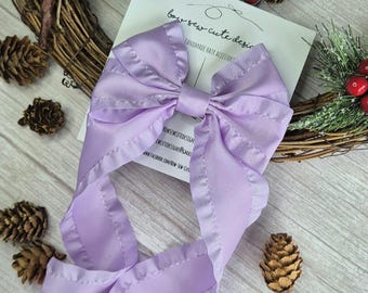 May include: A lavender satin bow with ruffled edges. The bow is tied with a long ribbon and is handmade. The text "bow-sew-cute designs" and "handmade hair accessories" is visible on a white card behind the bow.