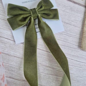 May include: Olive green velvet hair bow with long streamers. The bow is on a white card with the text "bow bow cute designs HANDMADE HAIR ACCESSORIES". The bow is on a light wood surface.