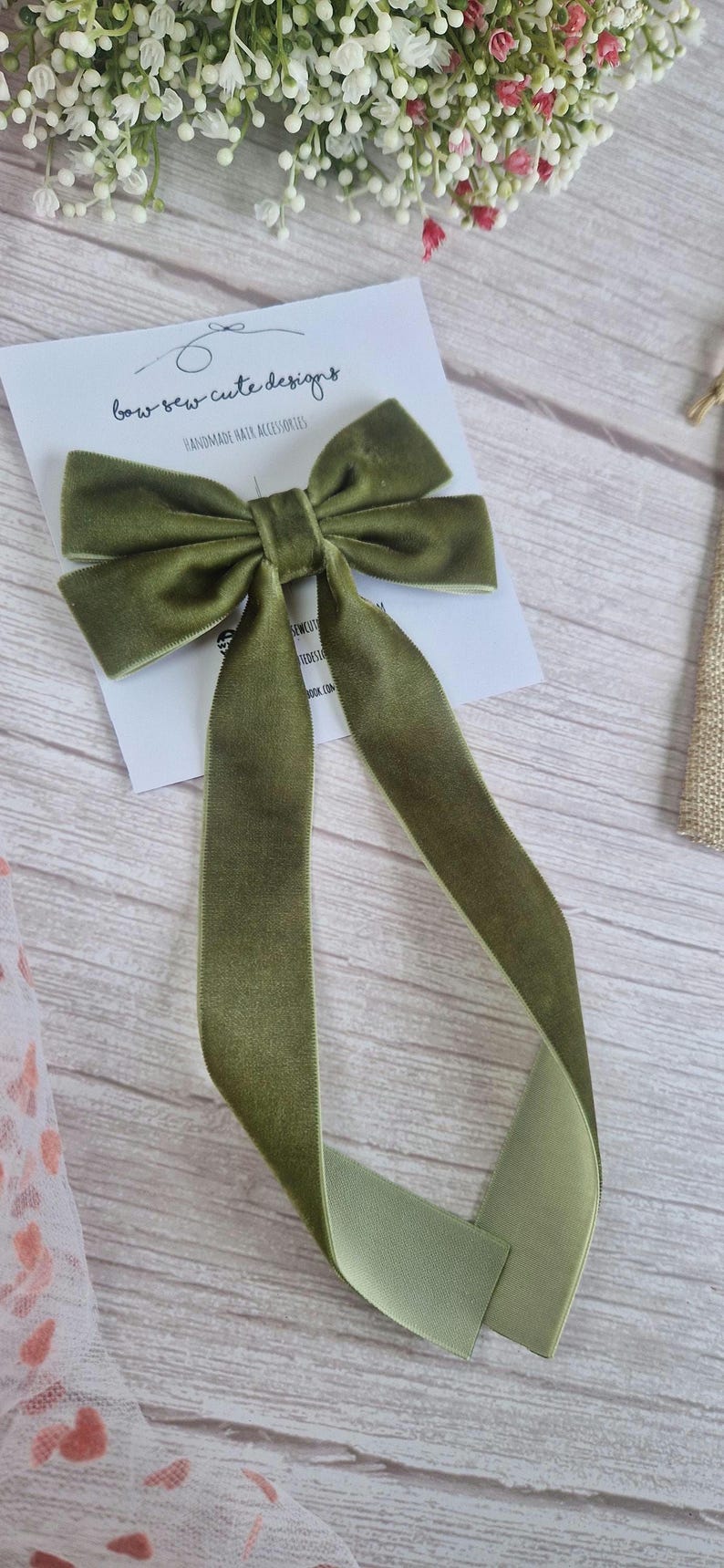May include: Olive green velvet hair bow with long streamers. The bow is on a white card with the text "bow bow cute designs HANDMADE HAIR ACCESSORIES". The bow is on a light wood surface.