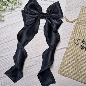 May include: Black satin hair bow with ruffled edges and long tails. The bow is tied with a small black elastic band. The bow is on a white wooden surface with a small burlap bag with a heart design.
