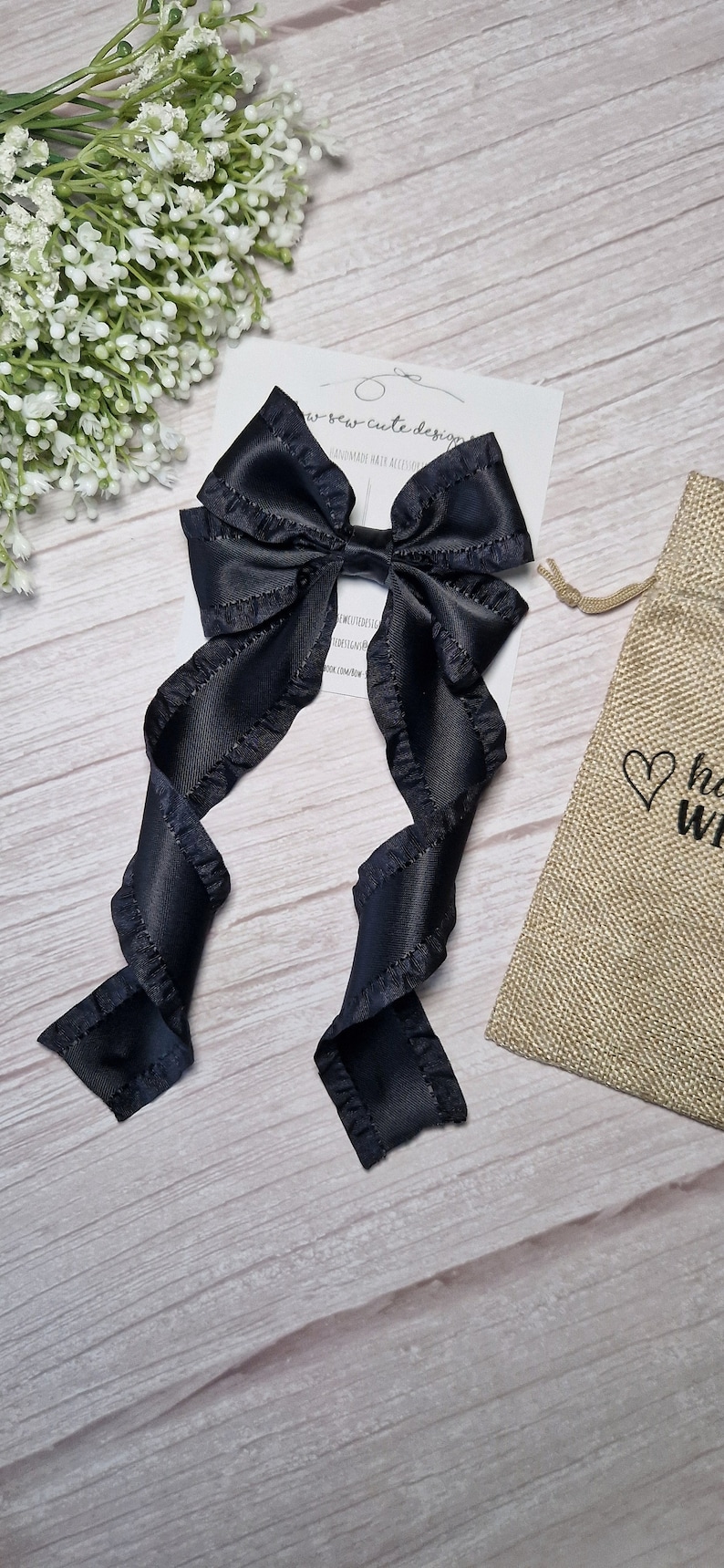 May include: Black satin hair bow with ruffled edges and long tails. The bow is tied with a small black elastic band. The bow is on a white wooden surface with a small burlap bag with a heart design.