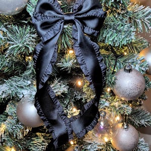 May include: A black satin bow with ruffled edges is tied around a flocked Christmas tree with silver ornaments and white lights.