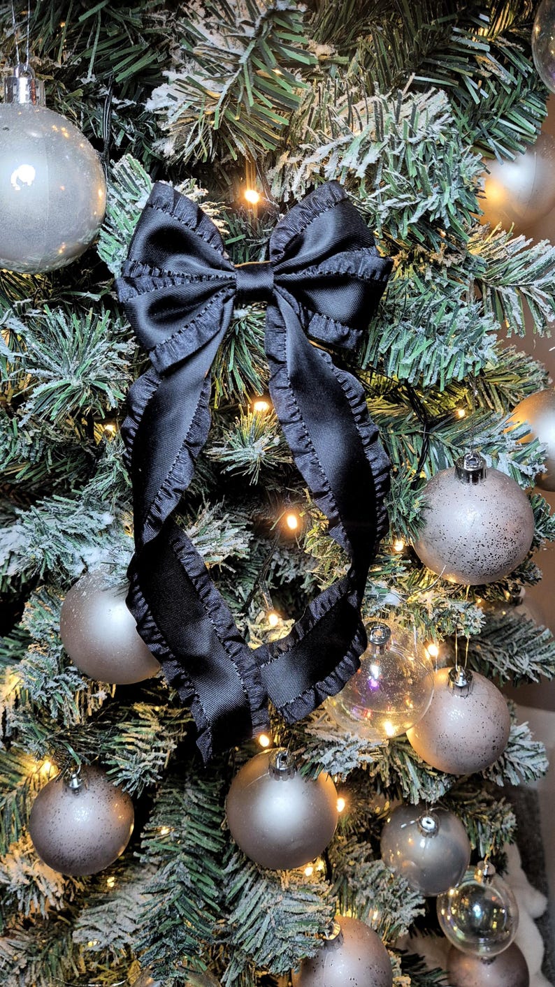 May include: A black satin bow with ruffled edges is tied around a flocked Christmas tree with silver ornaments and white lights.