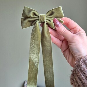 May include: A sage green velvet bow with long, flowing tails. The bow is held up against a neutral background. The fabric has a soft, textured appearance. This hair accessory is suitable for various occasions.
