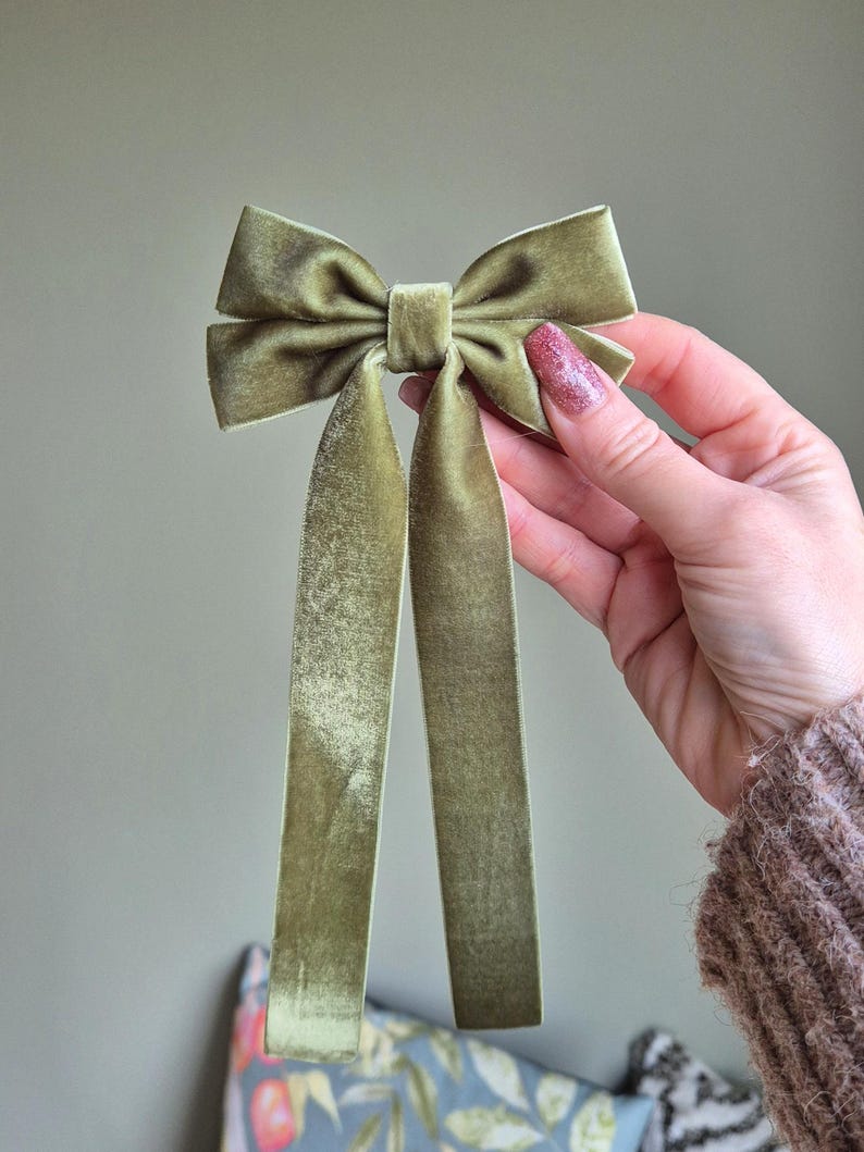 May include: A sage green velvet bow with long, flowing tails. The bow is held up against a neutral background. The fabric has a soft, textured appearance. This hair accessory is suitable for various occasions.