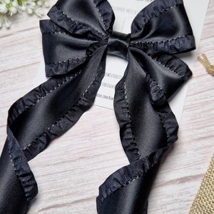 May include: A black satin ribbon bow with ruffled edges. The bow is tied with a long tail and is attached to a hair clip. The bow is on a white wooden surface.