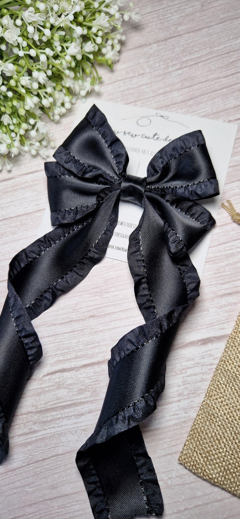 May include: A black satin ribbon bow with ruffled edges. The bow is tied with a long tail and is attached to a hair clip. The bow is on a white wooden surface.