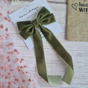 May include: An olive green velvet bow with long streamers, resting on a white card with the text "bow sew cute designs". A small burlap bag with a heart and the words "hand with" is visible in the background. Decorative hair accessory.