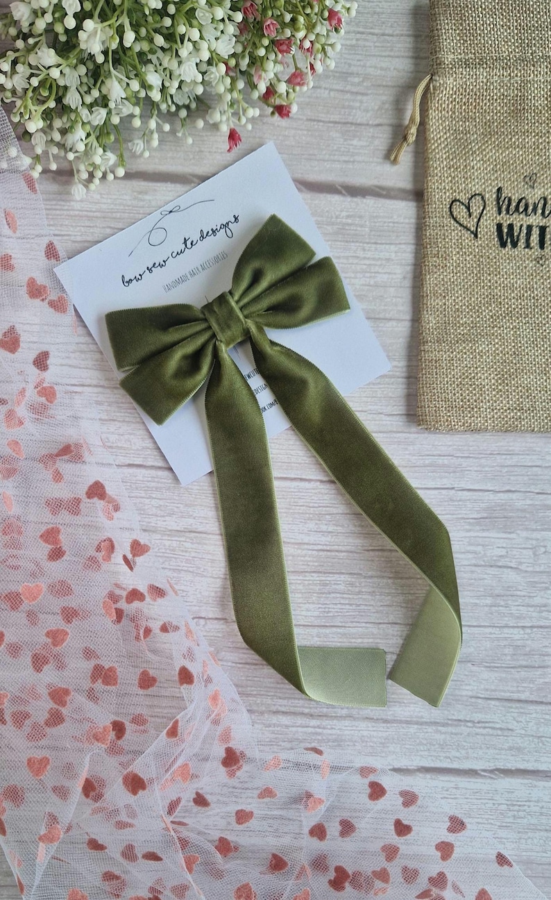 May include: An olive green velvet bow with long streamers, resting on a white card with the text "bow sew cute designs". A small burlap bag with a heart and the words "hand with" is visible in the background. Decorative hair accessory.