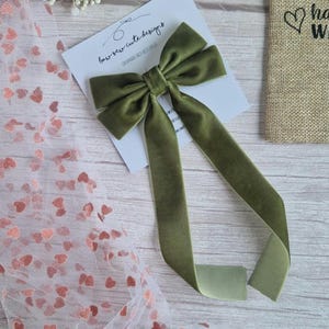 May include: Olive green velvet bow with long streamers, displayed on a white card with the text "bow sew cute designs". A small burlap bag with a heart graphic is visible in the background. Decorative accessory.
