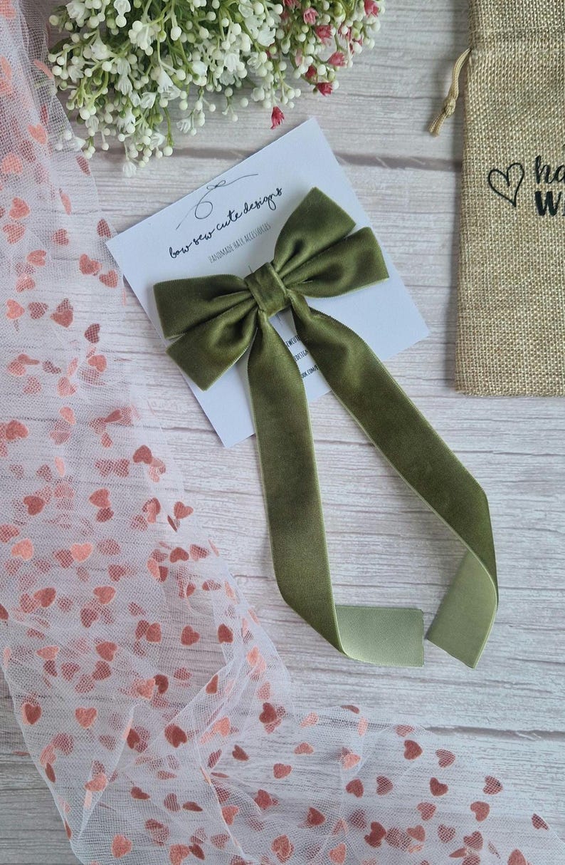 May include: Olive green velvet bow with long streamers, displayed on a white card with the text "bow sew cute designs". A small burlap bag with a heart graphic is visible in the background. Decorative accessory.