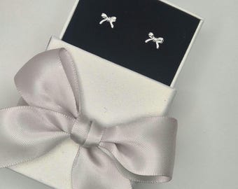 May include: A pair of silver bow-shaped stud earrings displayed in an open white gift box with a black interior. The box is tied with a large, light gray satin ribbon bow.