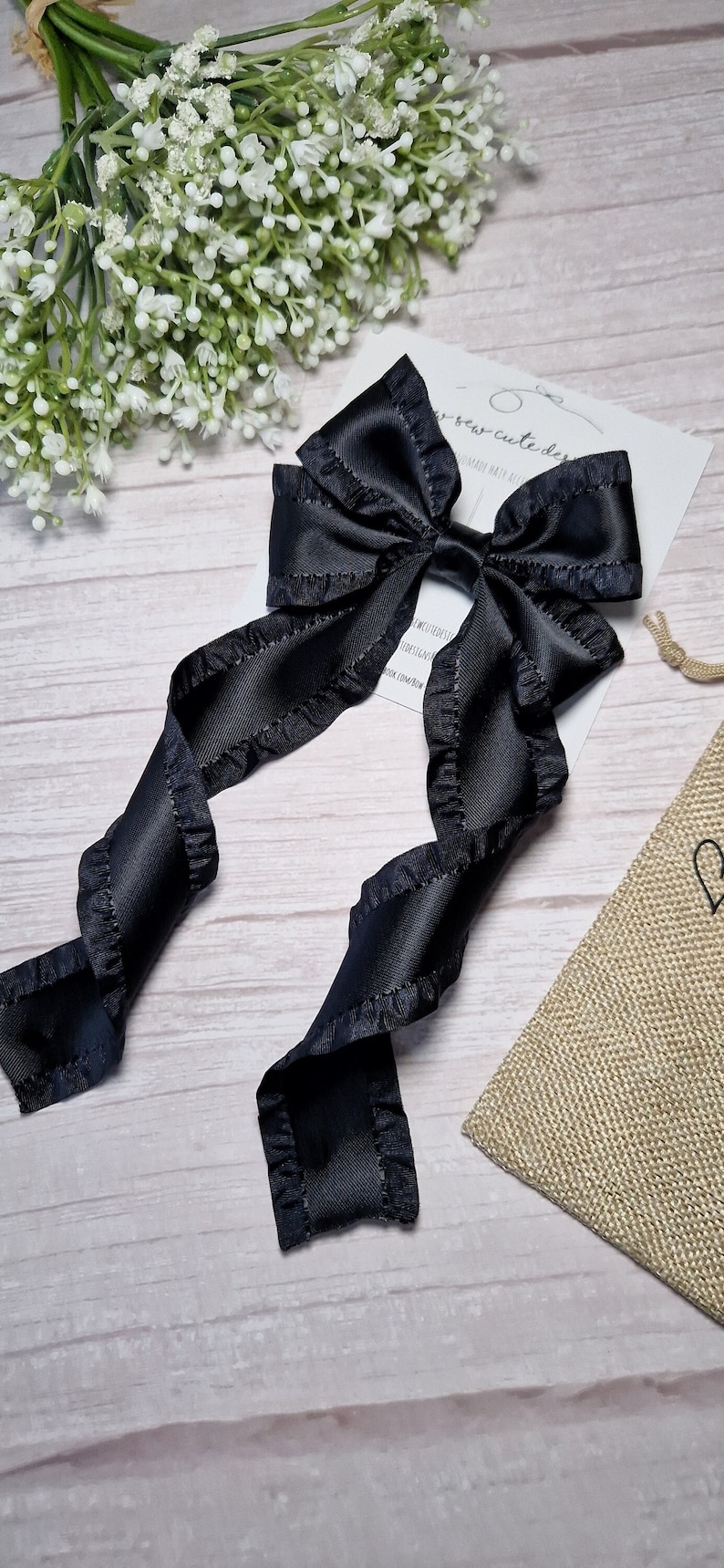 May include: A black satin ribbon bow with ruffled edges. The bow is tied with a long tail and is ready to be used as a hair accessory.