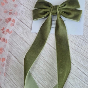 May include: Olive green velvet hair bow with long, flowing tails. The bow is tied in the center and sits atop a white card with the text "bow-sew cute designs" and "handmade hair accessories".