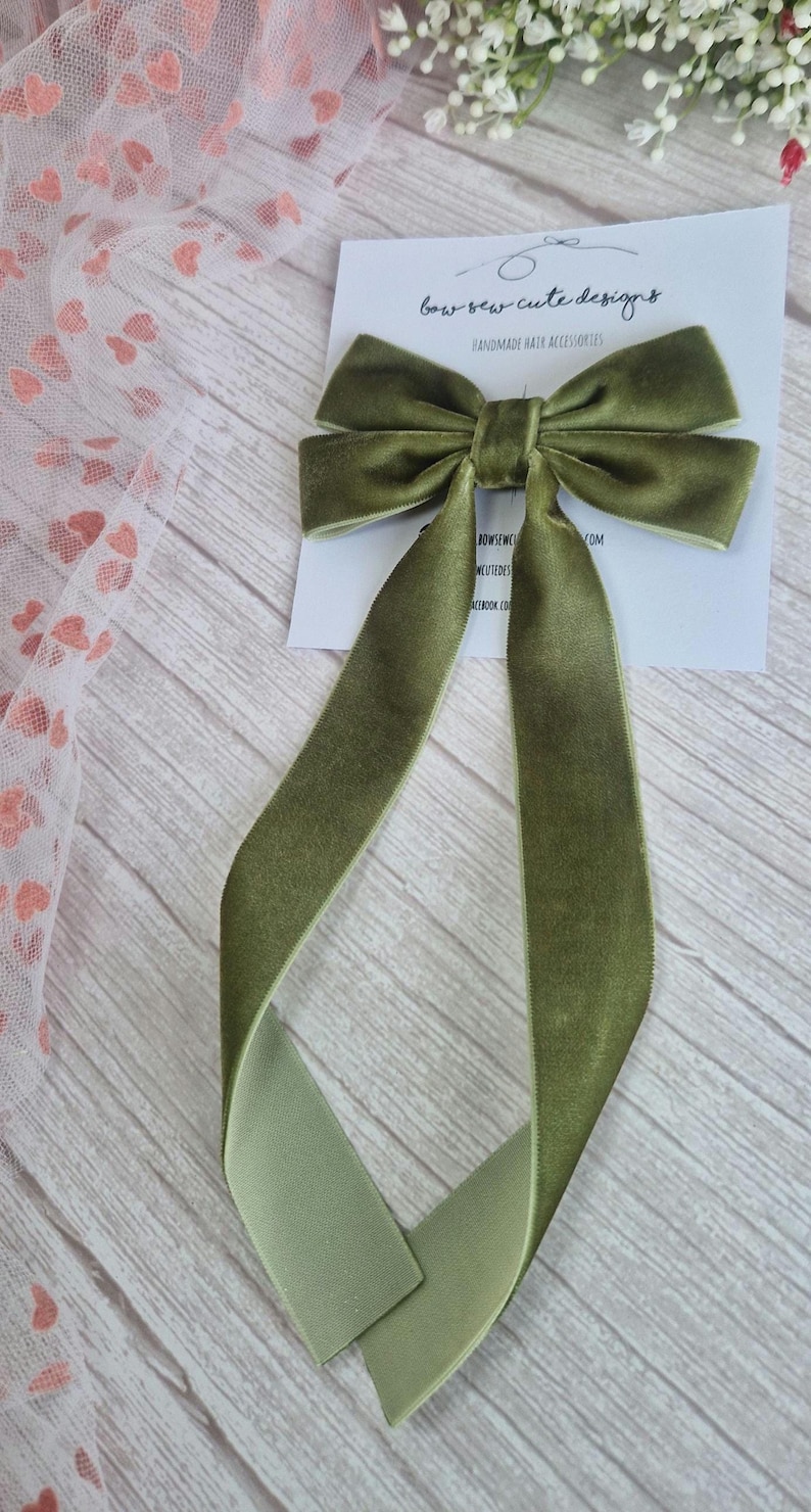 May include: Olive green velvet hair bow with long, flowing tails. The bow is tied in the center and sits atop a white card with the text "bow-sew cute designs" and "handmade hair accessories".