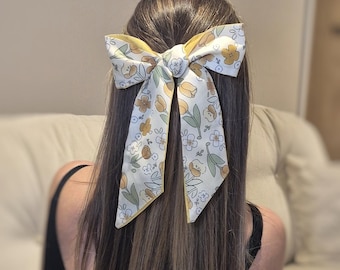 May include: A large hair bow with a floral pattern in yellow, white, and green is tied in the hair. The bow is set against a background of brown hair and a neutral-toned background.