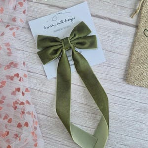 May include: Olive green velvet hair bow with long, flowing tails. The bow is centered on a white card with the text "bow-sew-cute designs". The background includes a sheer fabric with heart patterns and a wooden surface.