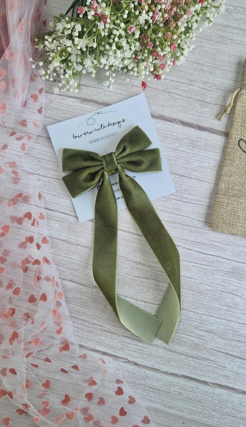 May include: Olive green velvet hair bow with long, flowing tails. The bow is centered on a white card with the text "bow-sew-cute designs". The background includes a sheer fabric with heart patterns and a wooden surface.