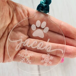 May include: Clear acrylic Christmas ornament with a paw print, the name "jacob" in cursive, and snowflake designs. It has a teal velvet ribbon for hanging. A festive and personalized holiday decoration.