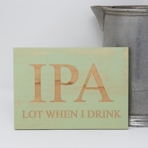May include: A green wooden sign with the text "IPA LOT WHEN I DRINK" in a light brown font. The sign is next to a silver pitcher.