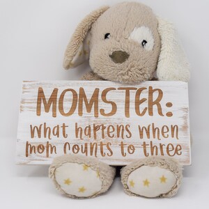 Puede incluir: Un letrero de madera blanco con el texto "MOMSTER: what happens when mom counts to three" en letras marrones. Un perro de peluche beige con orejas blancas está sosteniendo el letrero.