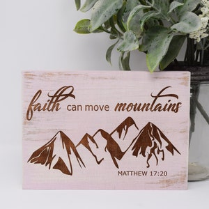 May include: A pink wooden sign with the text "faith can move mountains" and a silhouette of mountains. The text "MATTHEW 17:20" is printed below the mountains.