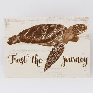 May include: A white wooden sign with a brown silhouette of a sea turtle and the text "Trust the journey".