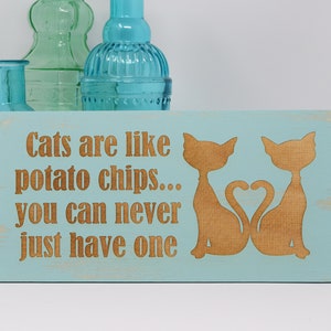 May include: A light blue wooden sign with the text "Cats are like potato chips... you can never just have one" in brown lettering. Two brown cat silhouettes are on the sign, facing each other with their tails forming a heart shape.